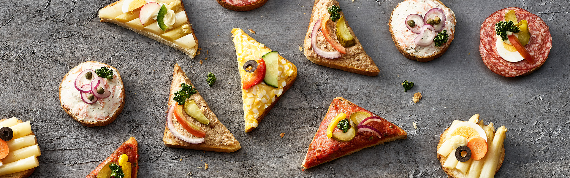 Dreieckige und runde Canapés der Sorten Lachs, Thon, Spargel, Ei, Tatar und Salami auf einem Stein präsentiert.