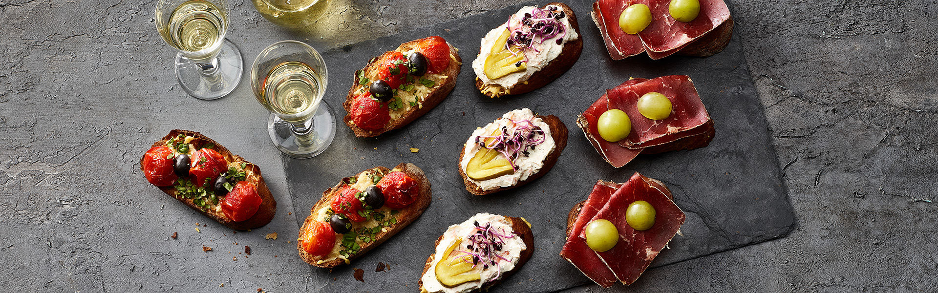 Header_Apéro.jpg Verschiedene Apéro Bruschetta sind auf einer Steinplatte präsentiert, dazu Weisswein in einer Karaffe.