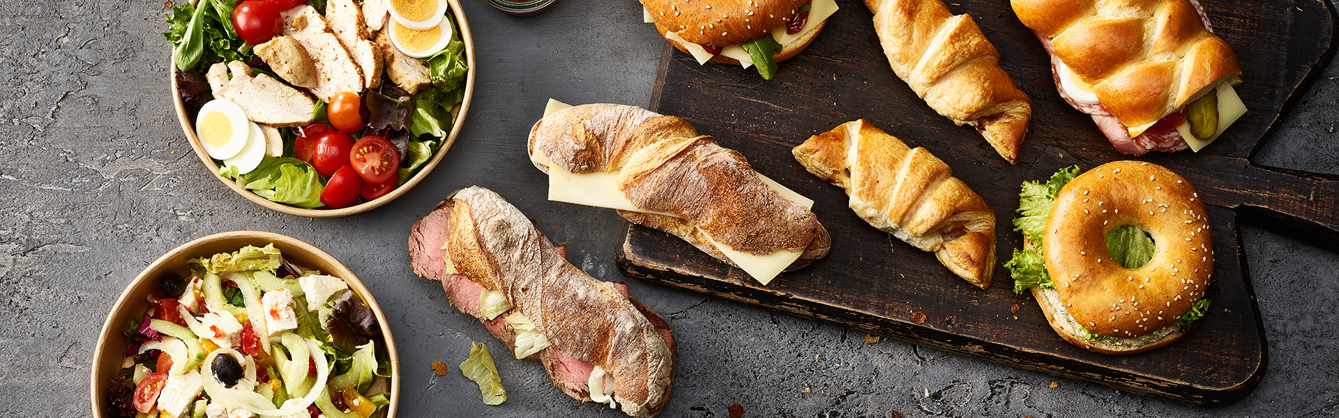Frische Salate, Sandwiches, Bagel und Schinkengipfeli liegen auf einem Holzbrett.