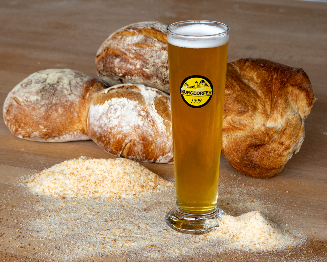Ein Glas Bäcker Bier steht in Paniermehl vor drei Laiben Brot.