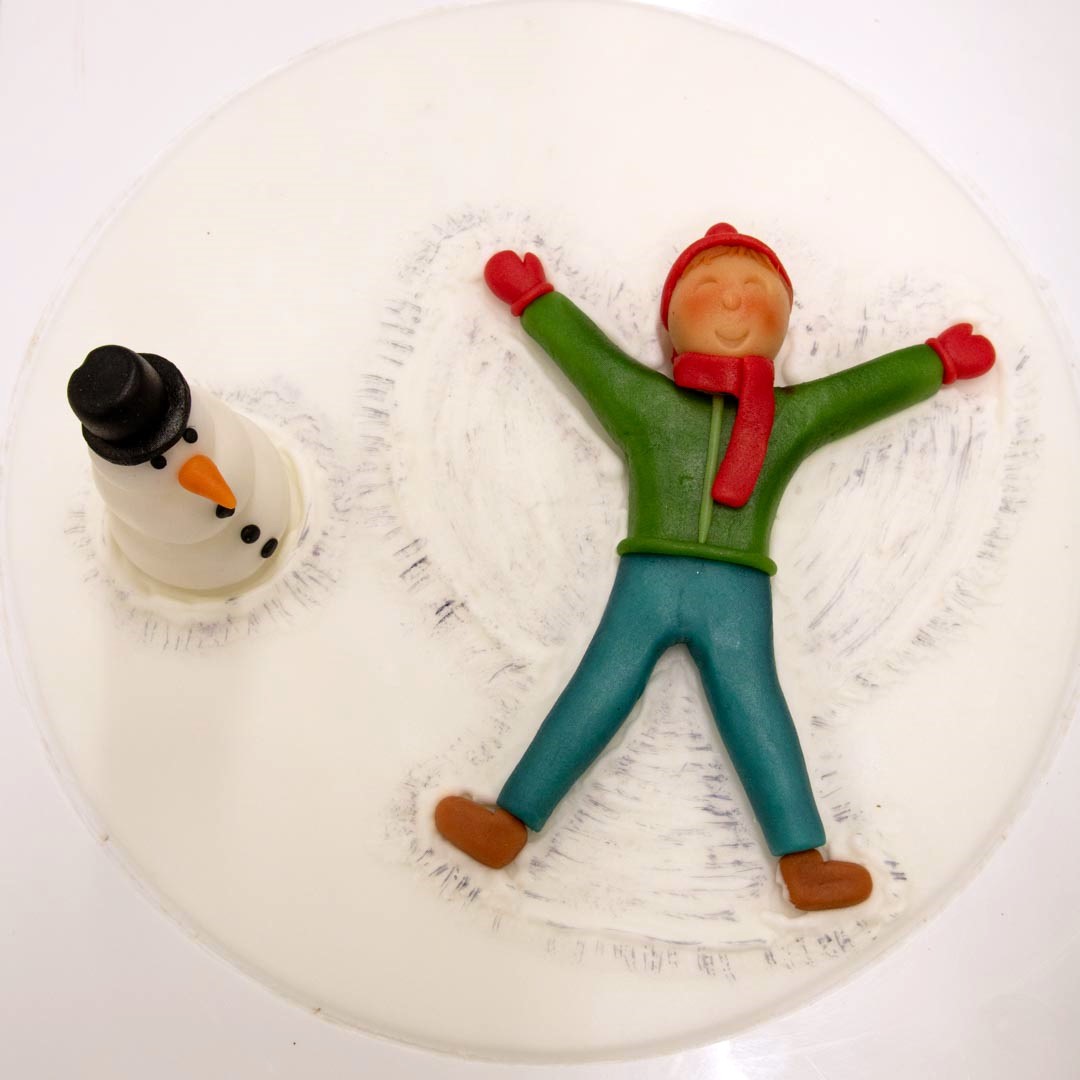 Runder Tortendecor mit Junge, der einen Schneeengel macht und daneben steht ein Schneemann aus Fondant.