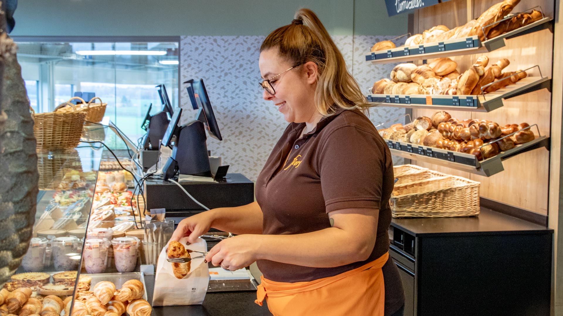 Mitarbeiterin packt ein Wienerli im Teig in einen Brotbeutel aus Papier, damit der Kunde es mitnehmen kann.