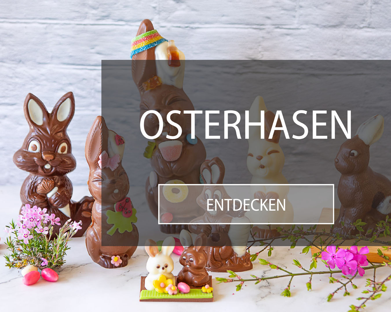 Osterhasen aus Schokolade mit verschiedenen Verzierungen stehen auf Marmoruntergrund, der mit Zuckereili und Blumen dekoriert ist.