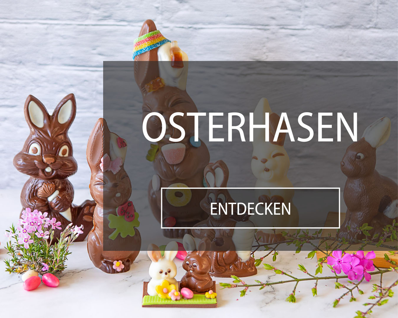 Osterhasen aus Schokolade mit verschiedenen Verzierungen stehen auf Marmoruntergrund, der mit Zuckereili und Blumen dekoriert ist.