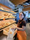 Jessica steht vor dem Brotgestell, lächelt in die Kamera und packt Brot in einen Brotsack.