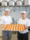 Tamina (links) und Maria (rechts) halten ein Blech gefüllt mit Wienerkonfekt in die Kamera und lächeln.